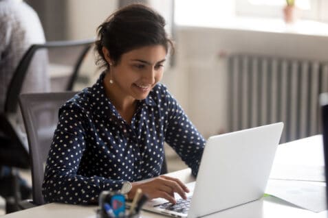 Smiling Indian Female Employee Using Laptop At Workplace